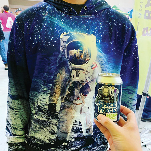 A person in an astronaut hoodie holds a beer from Hop & Barrel Brewing